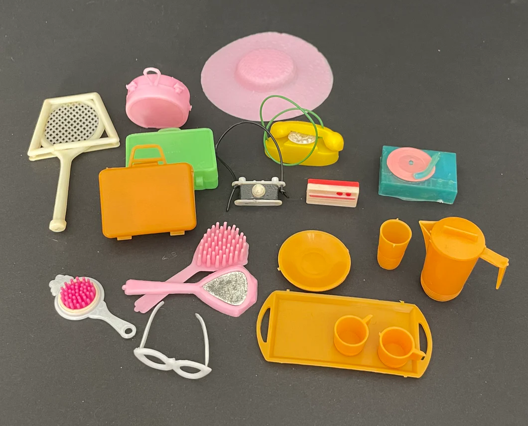 Barbie accessories from the 1970s, including a sunhat, sunglasses, water pitcher and cups, hairbrush and mirror, telephone, camera, record player, and tennis racket